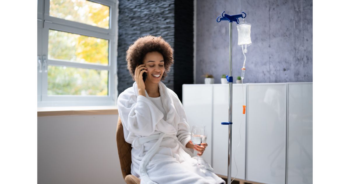 A person sitting comfortably in a robe while receiving an IV drip, smiling and talking on the phone, with a glass of water in hand, in a bright and cozy setting.