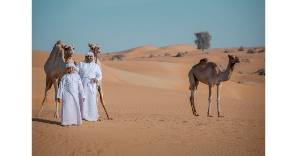 Two people in traditional attire standing with camels in the desert, with vast sand dunes in the background.
