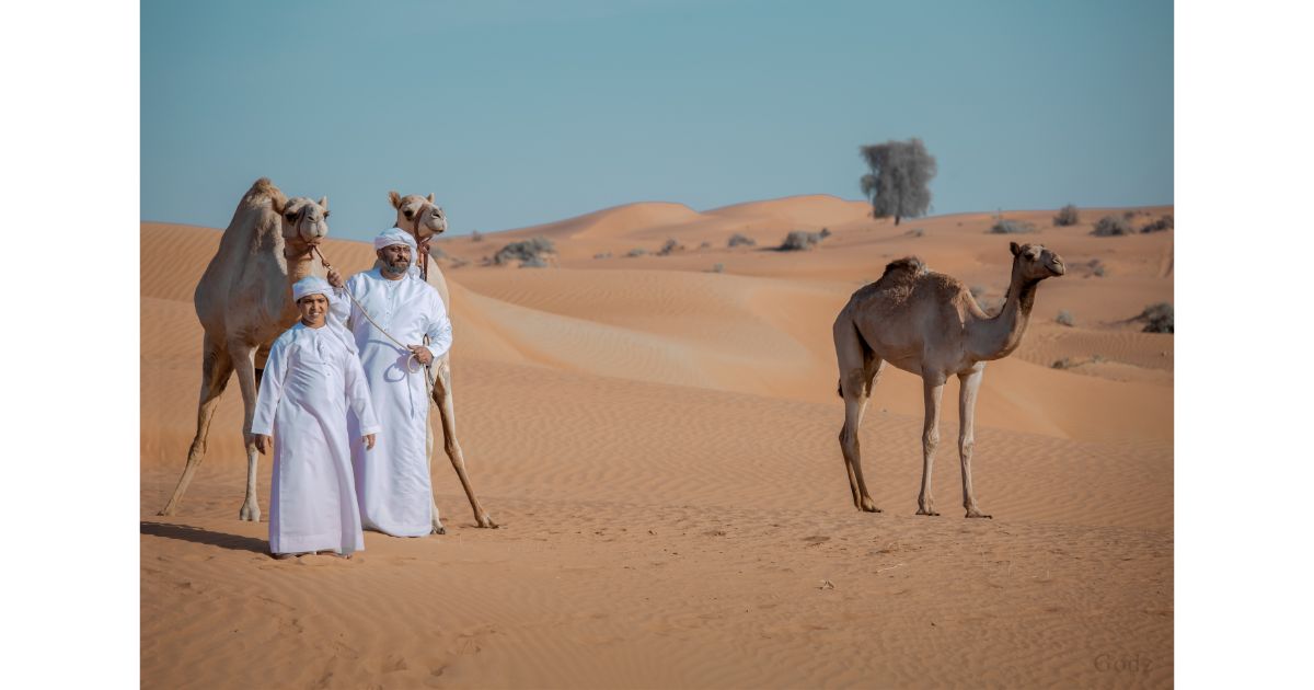 Two people in traditional attire standing with camels in the desert, with vast sand dunes in the background.