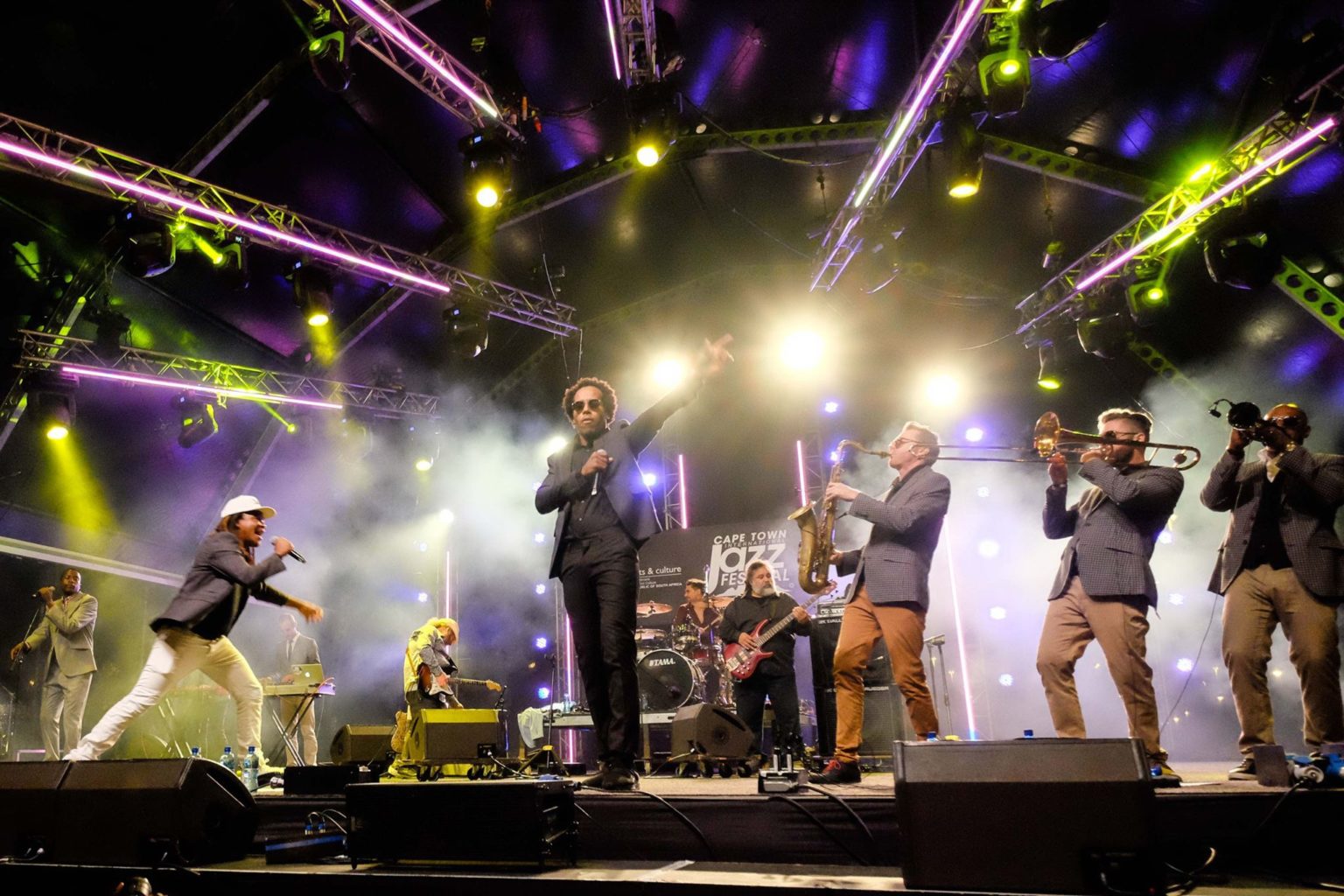 A lively jazz band performing on a brightly lit stage at a Cape Town music festival, with musicians playing saxophones, trumpets, and engaging the crowd.