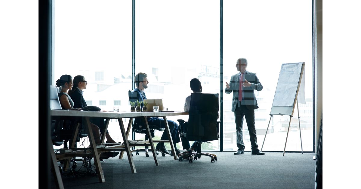 A business meeting in progress with a presenter speaking to a group of people sitting at a conference table, with a large window offering a city view in the background.