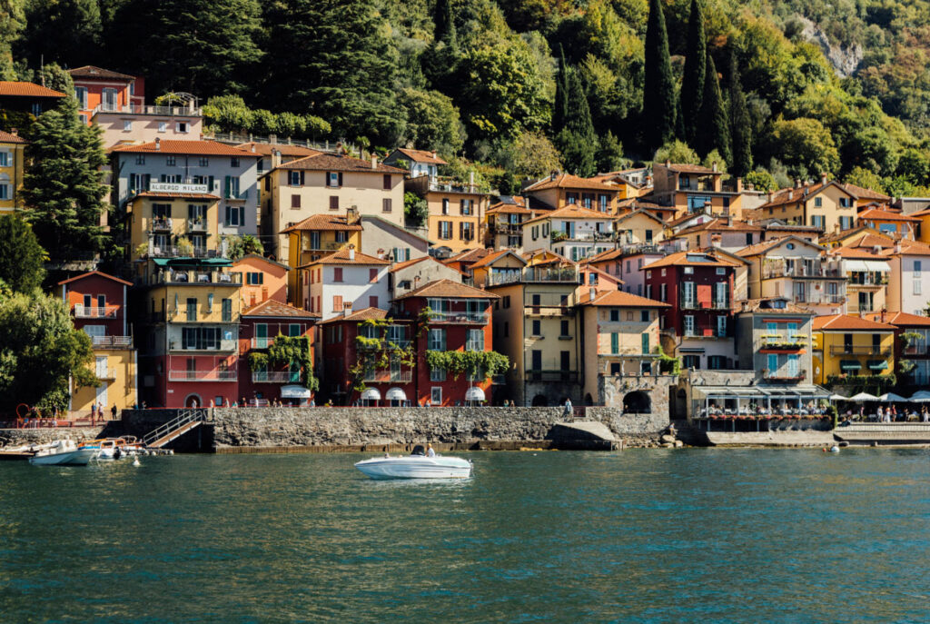A scenic view of colorful lakeside villas in Lake Como, Italy, with a white boat cruising on the calm waters, surrounded by lush greenery and mountains.