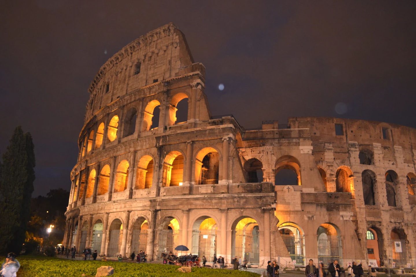The Colosseum in Rome illuminated at night, showcasing its ancient architecture with a dramatic evening sky in the background and visitors exploring the site.