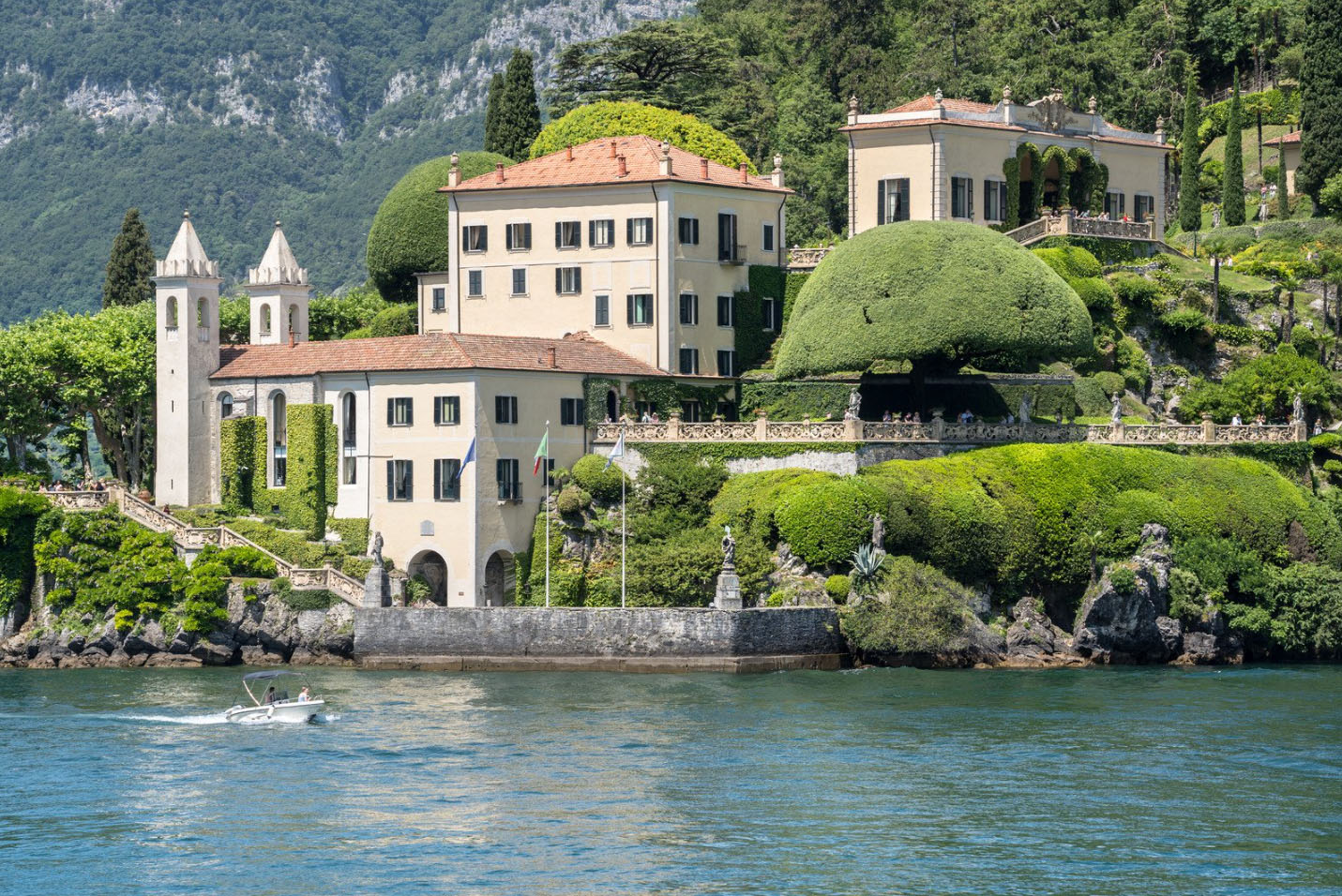 A stunning lakeside villa in Lake Como, Italy, surrounded by lush greenery and manicured gardens, with a small boat cruising on the calm waters.