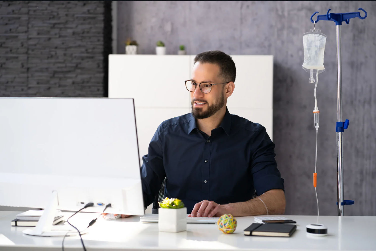 A smiling man working at his desk with an IV drip attached to his arm, indicating wellness and hydration therapy while staying productive.
