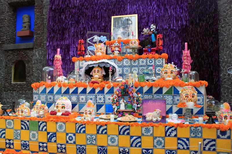 An altar for Día de los Muertos decorated with vibrant orange marigolds, colorful sugar skulls, candles, and other traditional offerings, set against a purple backdrop.