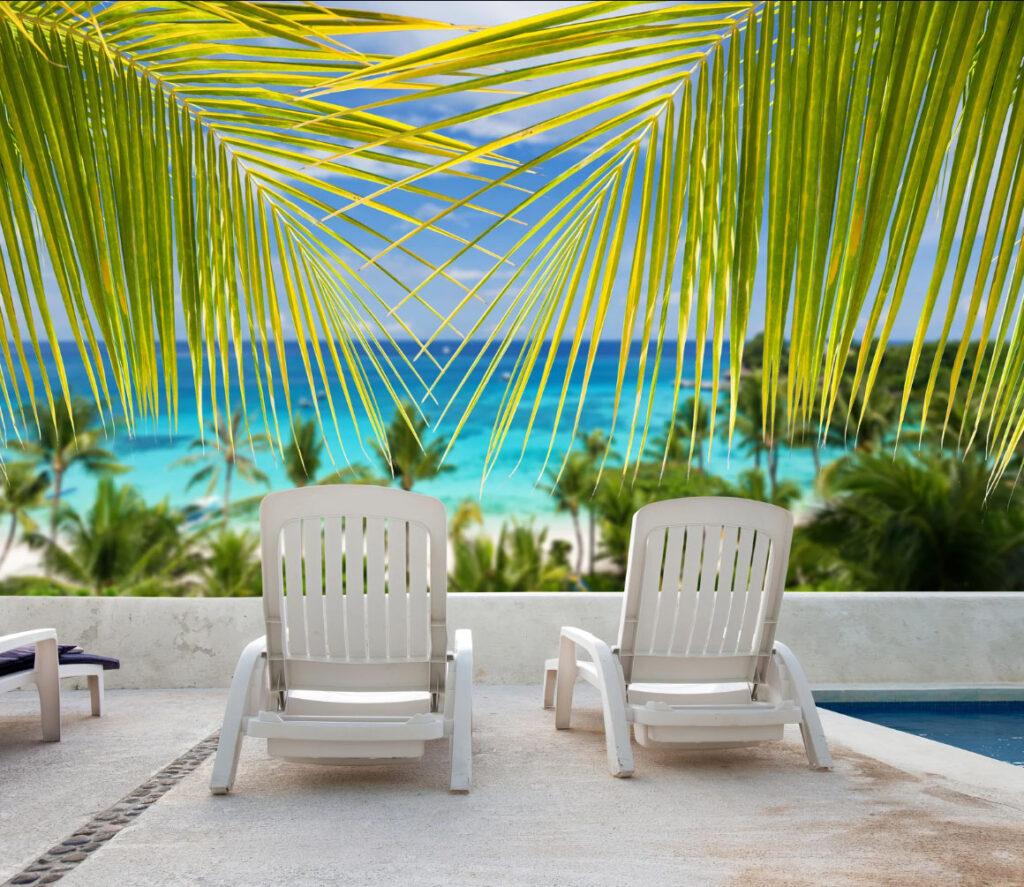 Two white lounge chairs on a tropical resort patio overlooking a turquoise ocean, framed by green palm leaves.