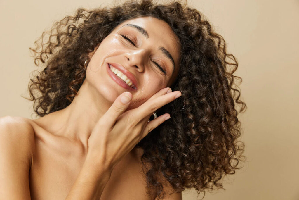A smiling woman with glowing, healthy skin and curly hair touching her face, radiating confidence and beauty.