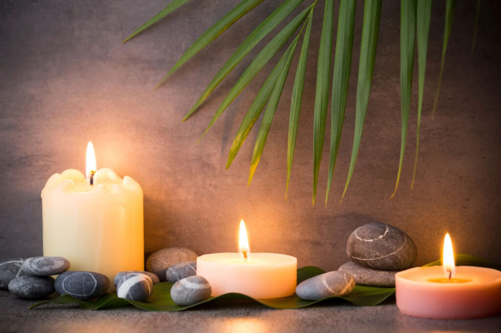 A serene spa setup featuring three lit candles, smooth gray stones, and green palm leaves against a dark, calming background.