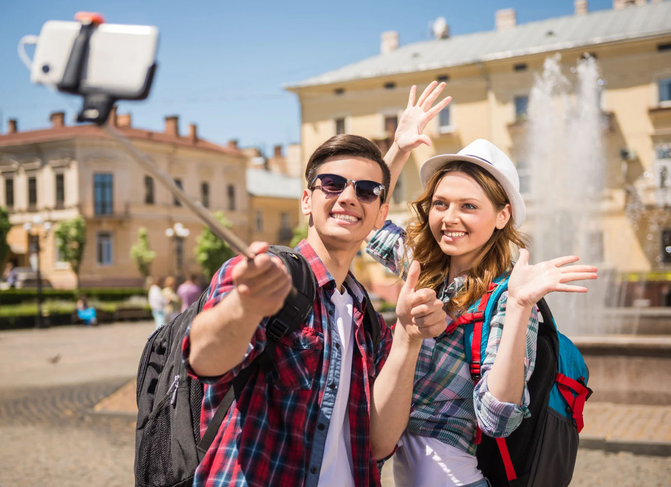 A young couple wearing backpacks and plaid shirts taking a selfie with a smartphone on a selfie stick in front of a fountain in a historic city square. They are smiling and waving, enjoying their travels.