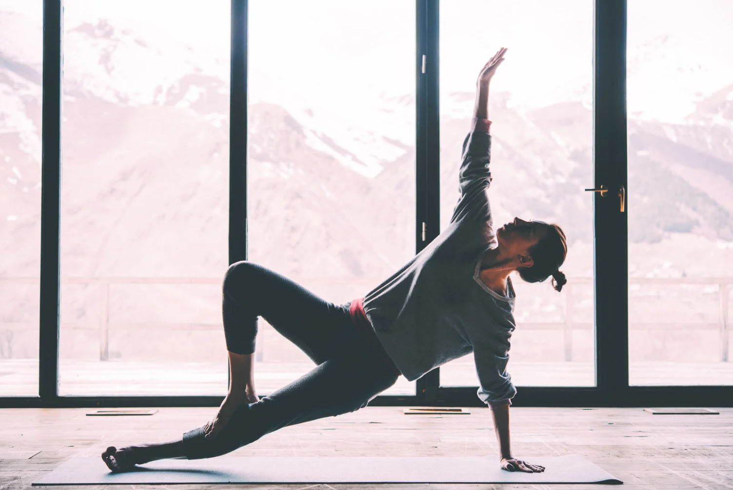 Woman practicing yoga indoors with a scenic mountain view in the background, performing a side plank pose on a yoga mat.