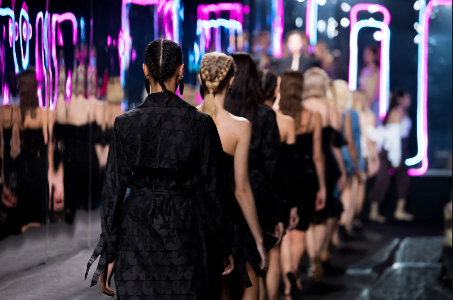 Models walking down the runway at Milan Fashion Week, showcasing high-end fashion against a backdrop of neon lights and a stylish audience.