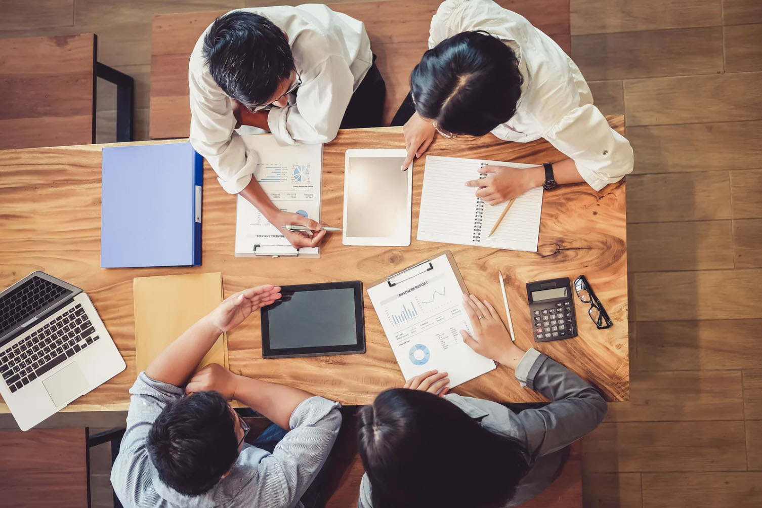 Overhead view of four people sitting around a wooden table, collaborating with documents, tablets, a notebook, and a calculator.