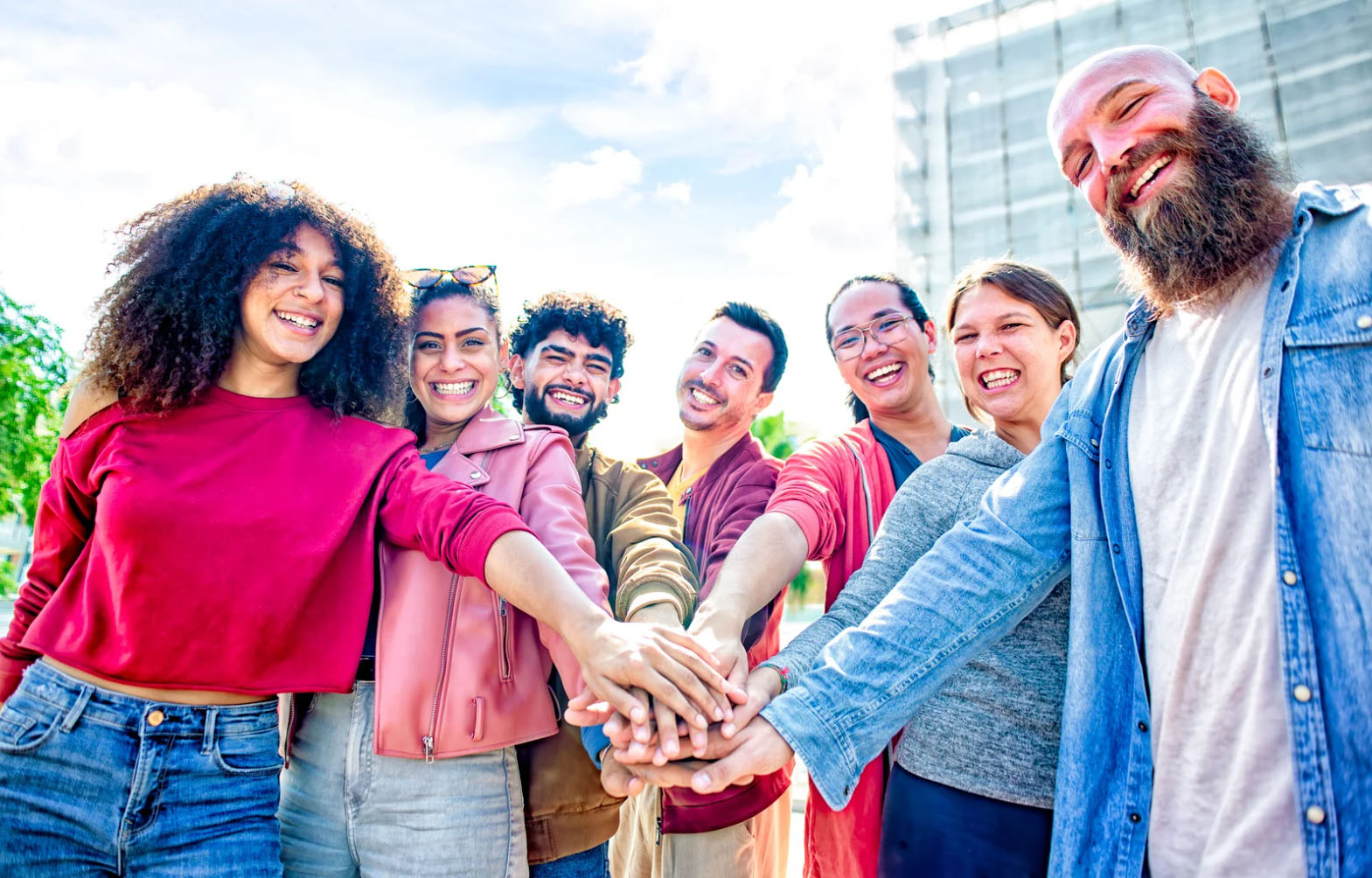 Group of diverse, smiling people standing in a row outdoors with their hands stacked together in a gesture of unity and teamwork.