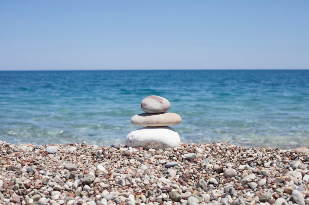 Stack of smooth stones balanced on a pebble beach with the ocean and clear blue sky in the background.