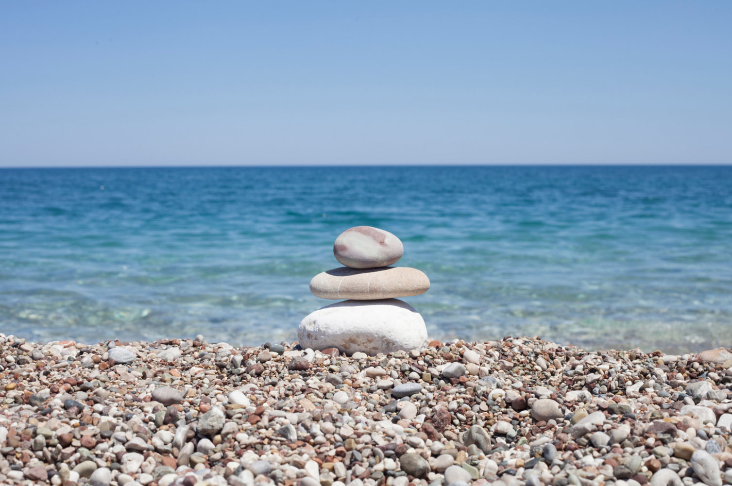 Stack of smooth stones balanced on a pebble beach with the ocean and clear blue sky in the background.