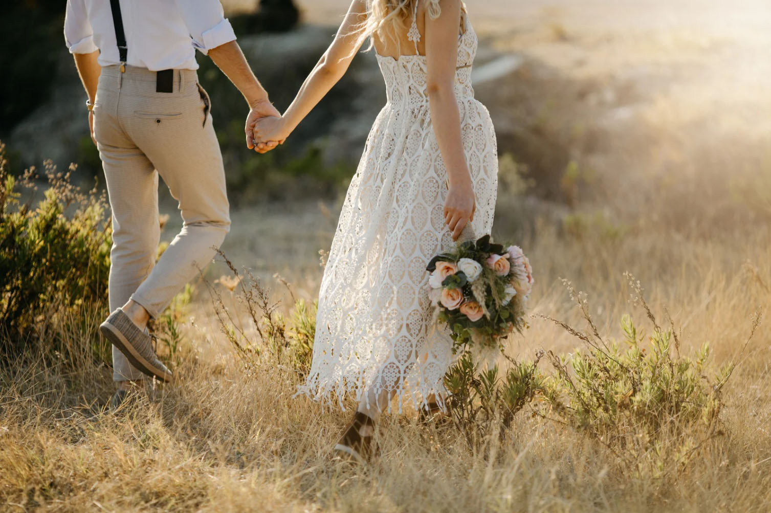 A couple holding hands while walking through a grassy field. The woman is wearing a lace wedding dress and holding a bouquet of pink roses, while the man is dressed in a white shirt, beige pants, and suspenders.