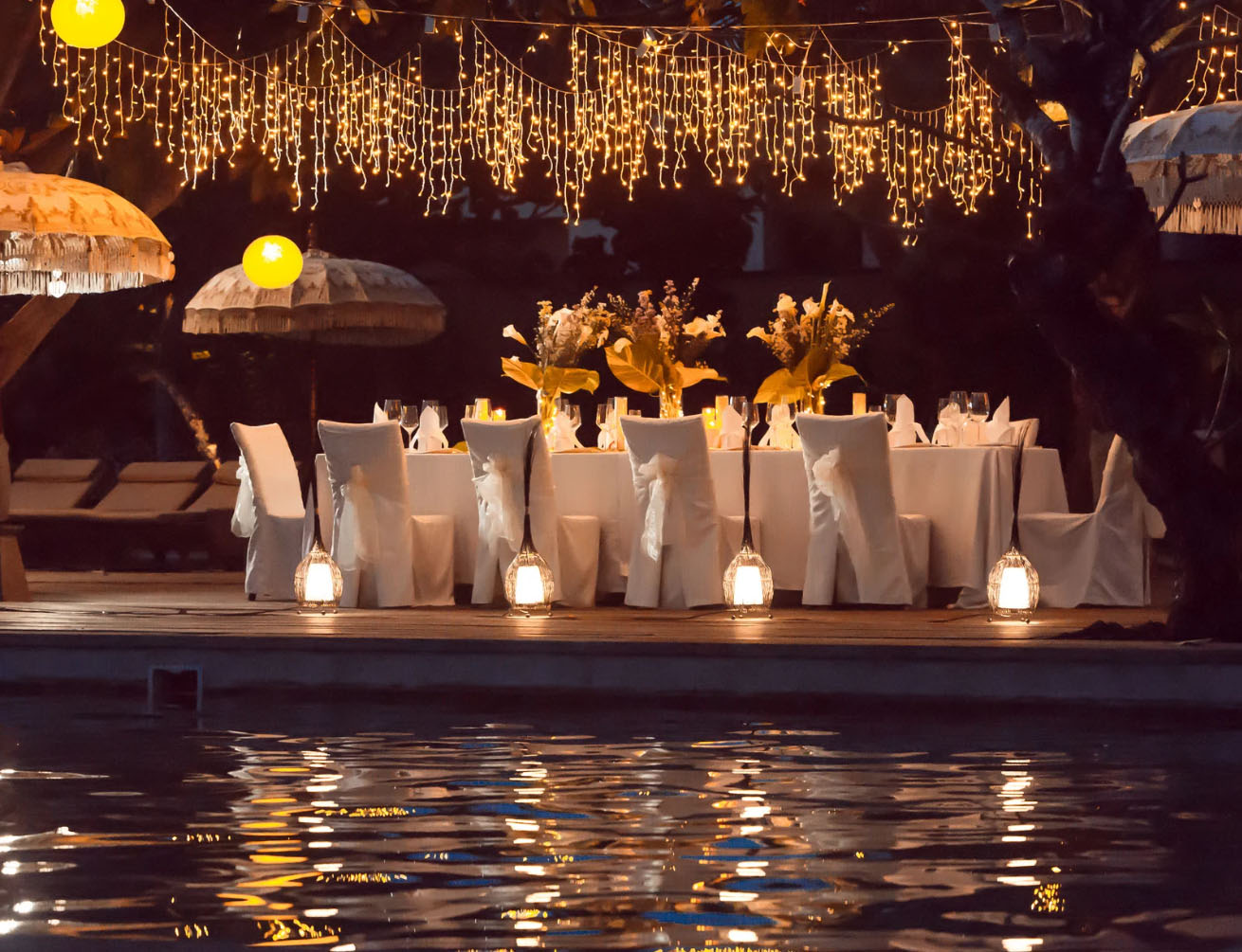 An elegantly set outdoor dining table under a canopy of twinkling fairy lights, with white chairs draped in fabric and lanterns around the table. The scene is reflected in the calm pool water.