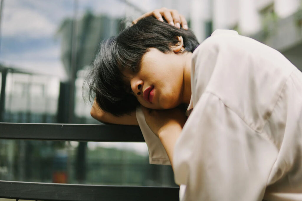 A person with short black hair leans on a glass railing, wearing a white shirt, looking down in a relaxed pose.