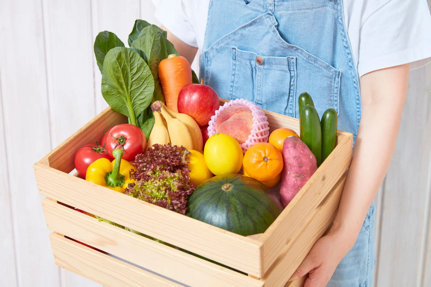 A person holding a wooden crate filled with a variety of fresh vegetables and fruits, including tomatoes, cucumbers, carrots, bananas, leafy greens, sweet potatoes, and watermelon.