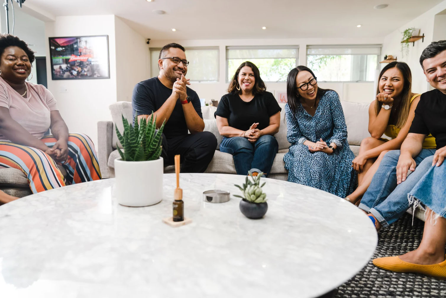 A group of six people seated around a coffee table in a cozy, modern living room. They are smiling and enjoying each other's company, with some engaging in conversation and others laughing. The room has natural light coming through large windows and a few plants on the table.
