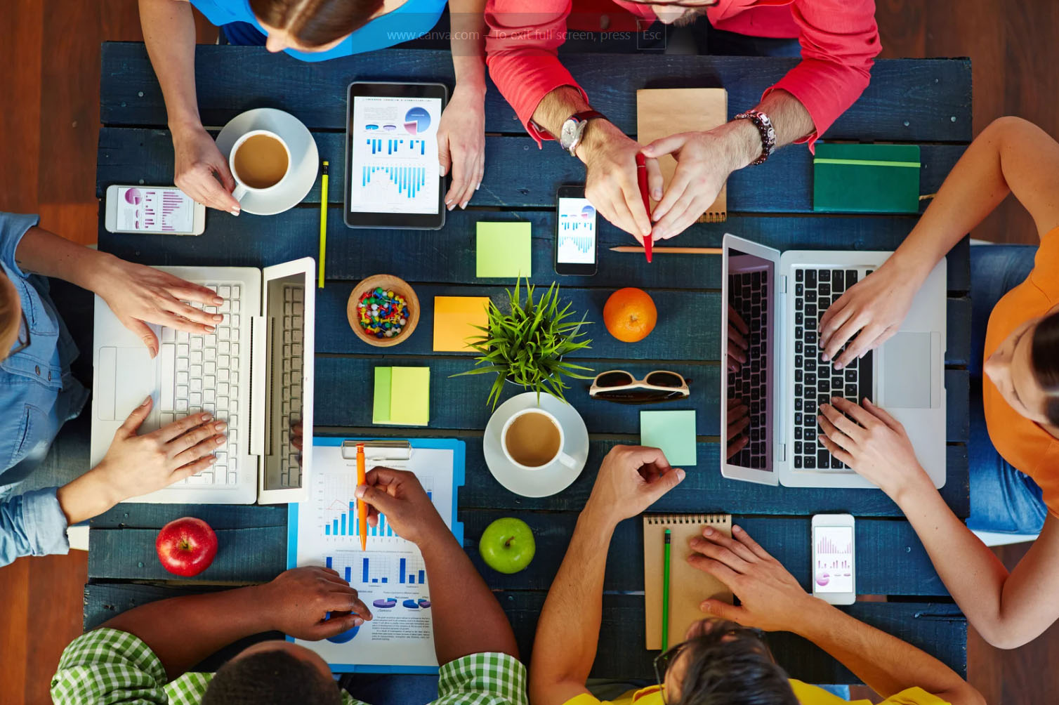 A top-down view of a group of people working together at a table with laptops, tablets, coffee cups, and colorful stationery items. There are also fruits like apples and oranges placed on the table, and people are engaged in discussions or taking notes.