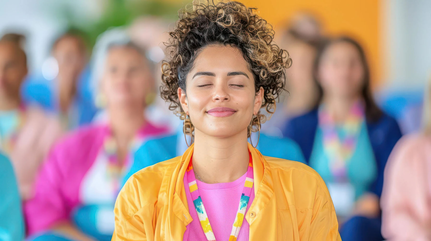 A woman with curly hair is smiling and meditating with her eyes closed in a group setting, wearing a yellow jacket, pink shirt, and a multicolored lanyard. The background shows a crowd of people blurred out.