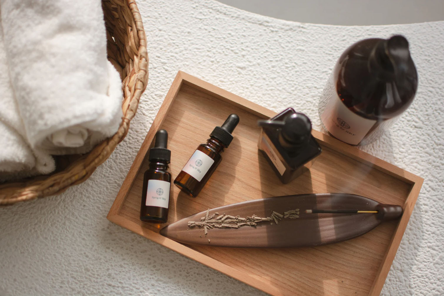 A wooden tray with essential oil bottles, a decorative incense holder with a leaf design, and neatly rolled towels arranged on a white surface.