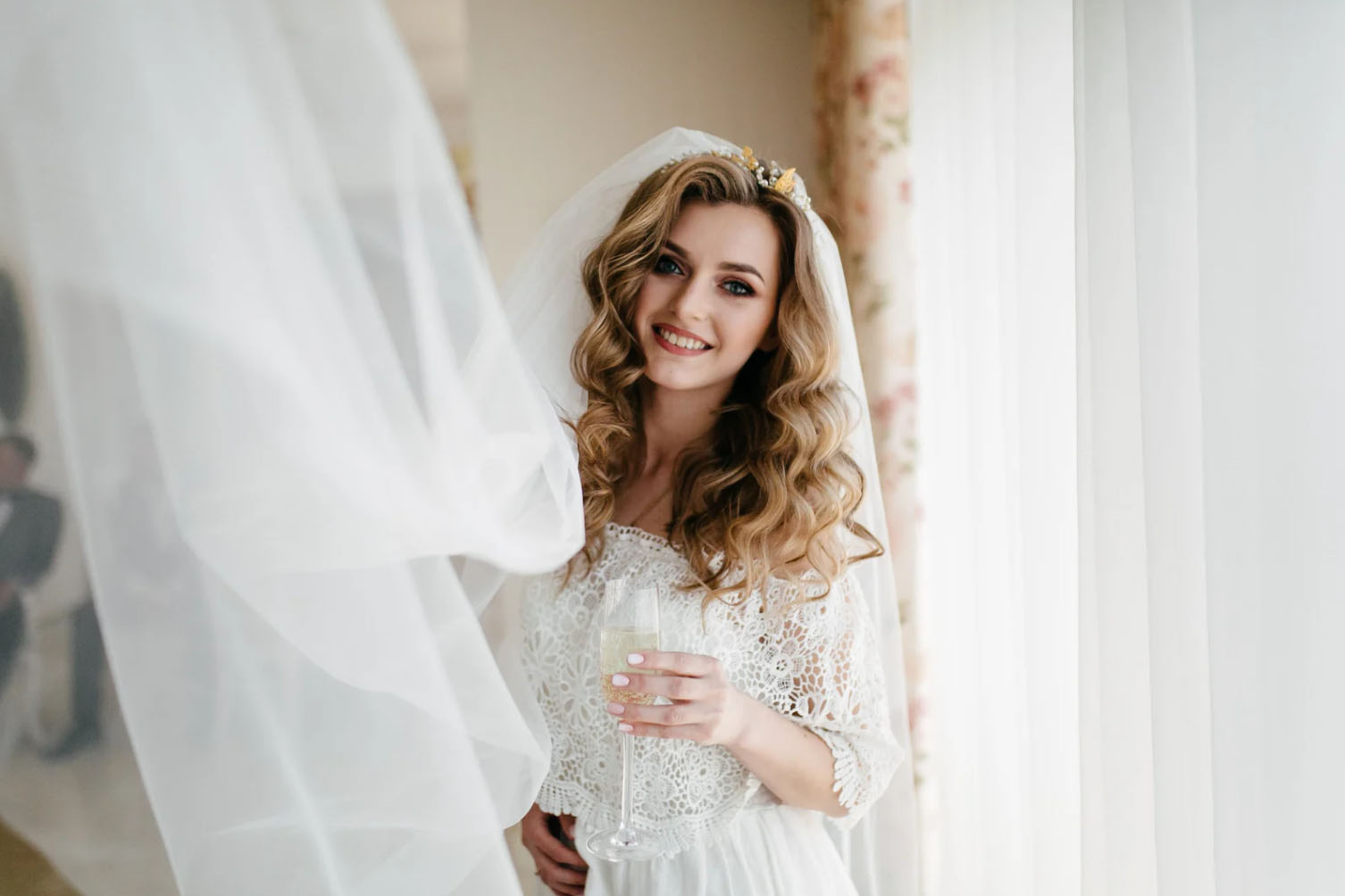 A bride with wavy hair and a floral crown smiles while holding a glass of champagne. She is surrounded by flowing wedding veil fabric and a softly lit window, giving the scene a dreamy atmosphere.