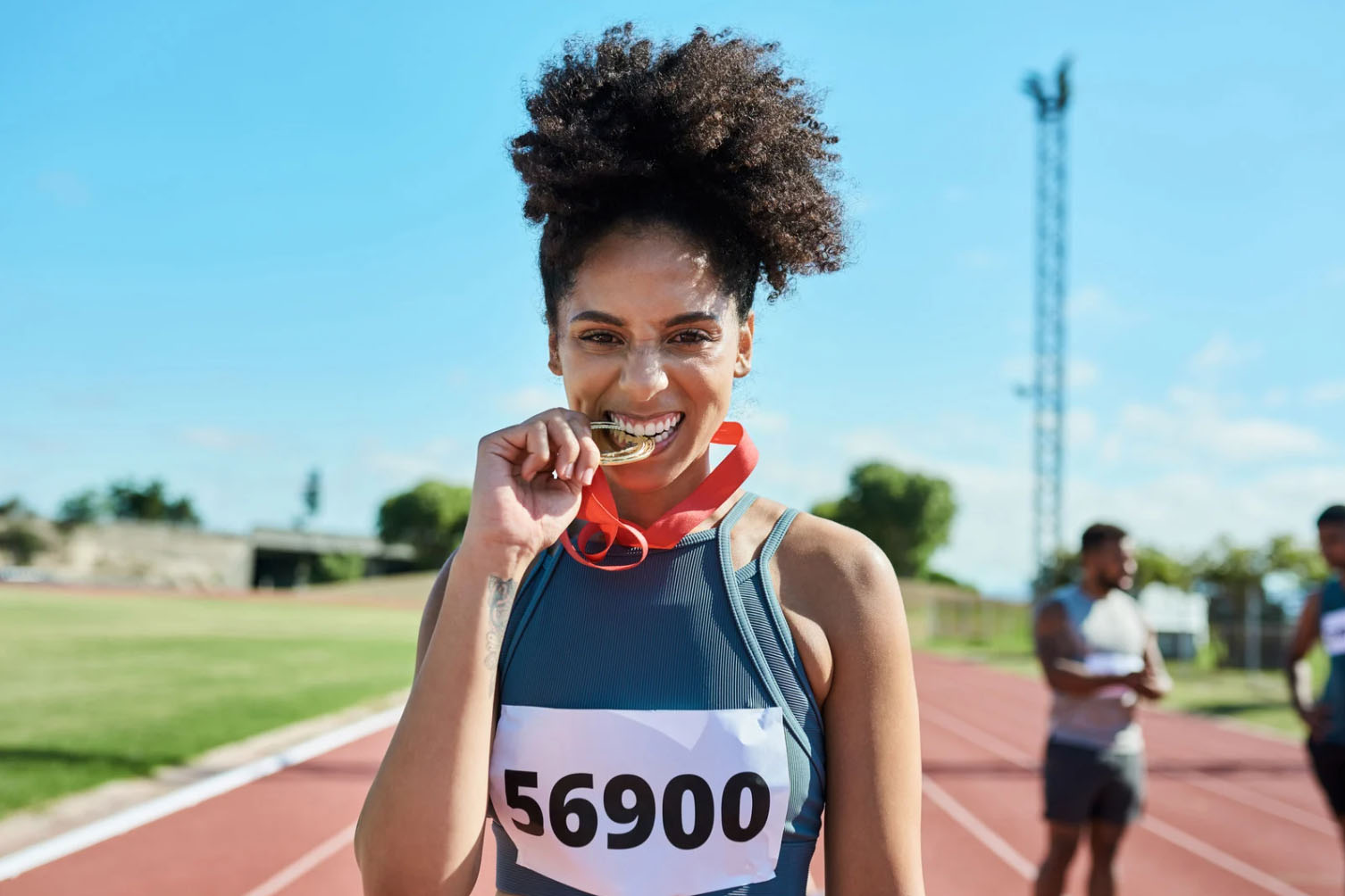 A smiling female athlete wearing a numbered bib and a red ribbon around her neck, holding a gold medal in her teeth, standing on a running track with other people in the background.