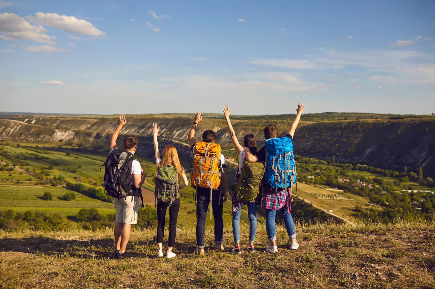 Group of friends with backpacks raising their hands while overlooking a scenic valley and mountains during a hike.