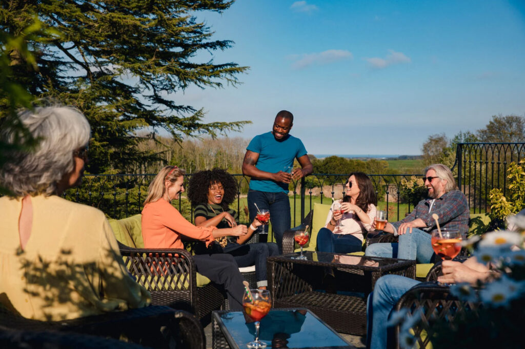 A group of six people, including both men and women, enjoy drinks and conversation outdoors on a sunny day. The group is seated on outdoor furniture, surrounded by greenery with a clear blue sky in the background.