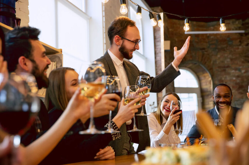 A group of people toasting with wine glasses, celebrating at a gathering in a cozy indoor setting. One person is standing and giving a speech, while others are smiling and enjoying the moment. The room has a warm atmosphere with string lights hanging above.