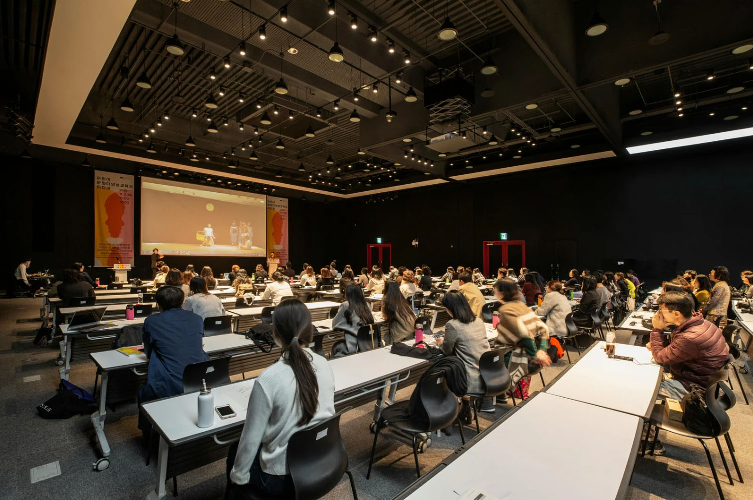 A large conference room filled with attendees seated at tables, watching a presentation on a screen. The room has modern lighting and a sleek design.