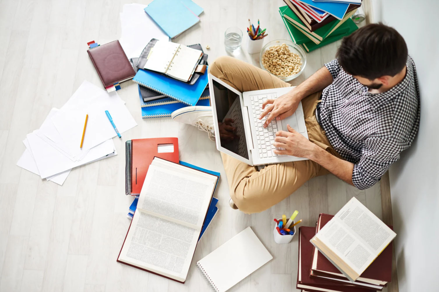 A man sitting on the floor surrounded by open books, notebooks, and papers, working on a laptop. The scene suggests studying or research, with a bowl of snacks and writing supplies nearby.
