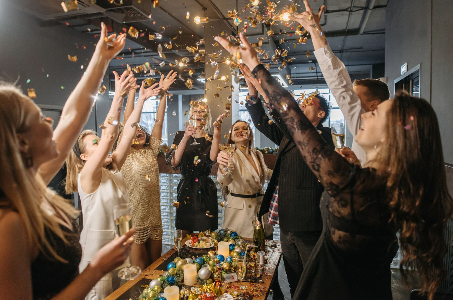 A group of people celebrating at a party, with confetti falling in the air. They are holding drinks and cheering, surrounded by a festive table with candles and decorations.