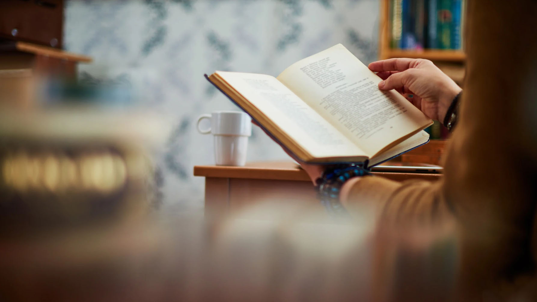 A person holding an open book, reading, with a cup placed on a nearby wooden surface. The background shows blurred shelves and wallpaper.