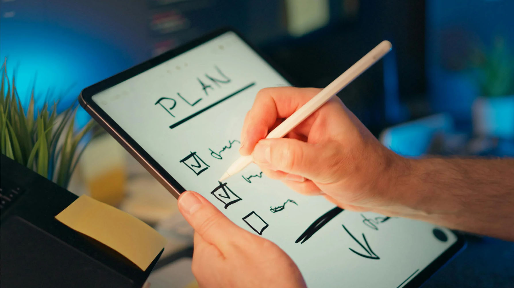 A person using a stylus on a tablet to check off items on a digital to-do list labeled 'PLAN,' with a workspace setting featuring plants and a yellow sticky note in the background.