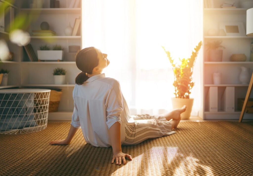 A person sitting on the floor in a sunlit room with a relaxed posture, facing away from the camera. They are wearing a light-colored shirt and striped pants, surrounded by plants and bookshelves in a cozy living space.