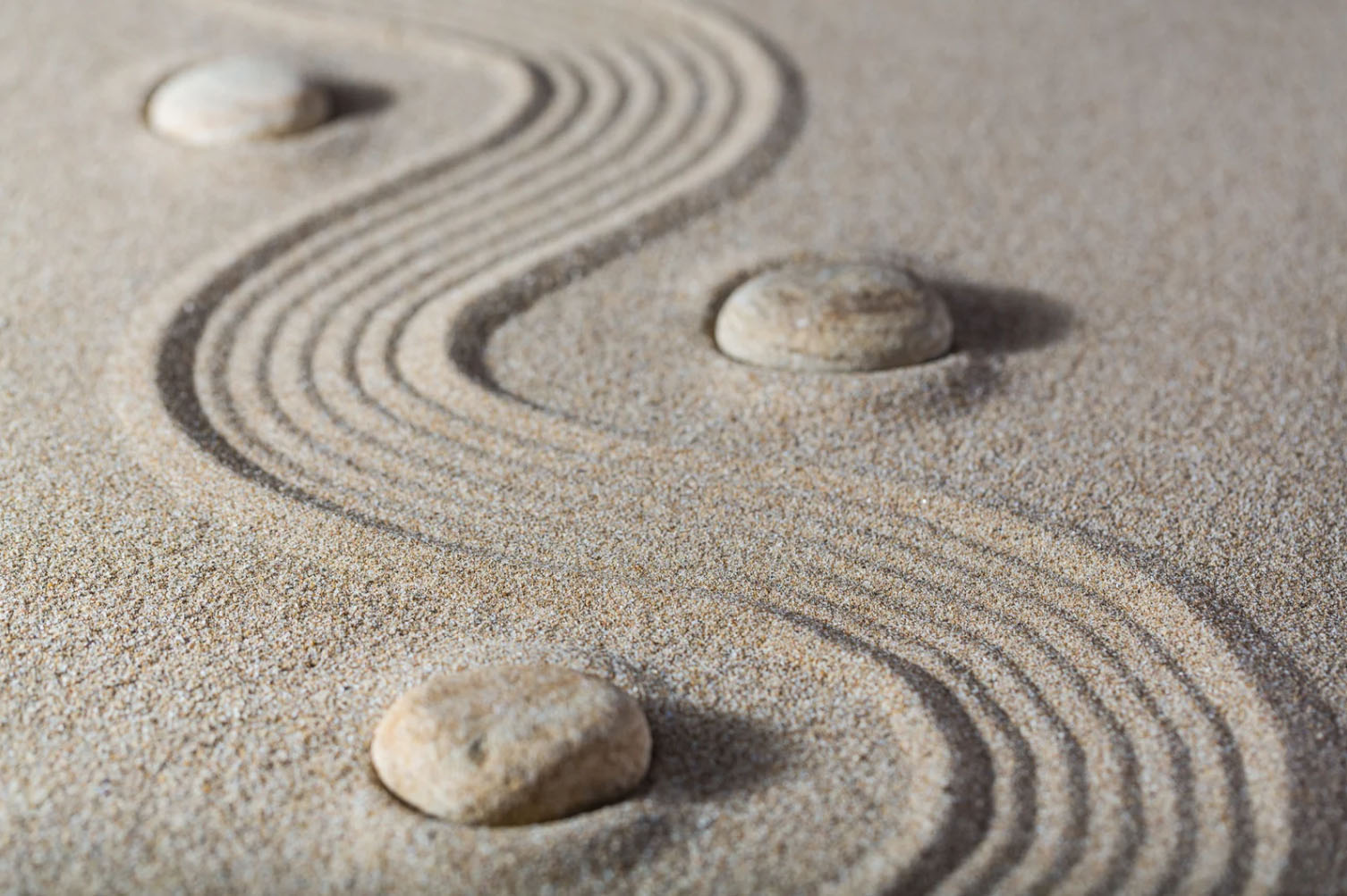A close-up view of smooth stones placed on a sandy surface, with concentric, wavy patterns gently etched in the sand around them.