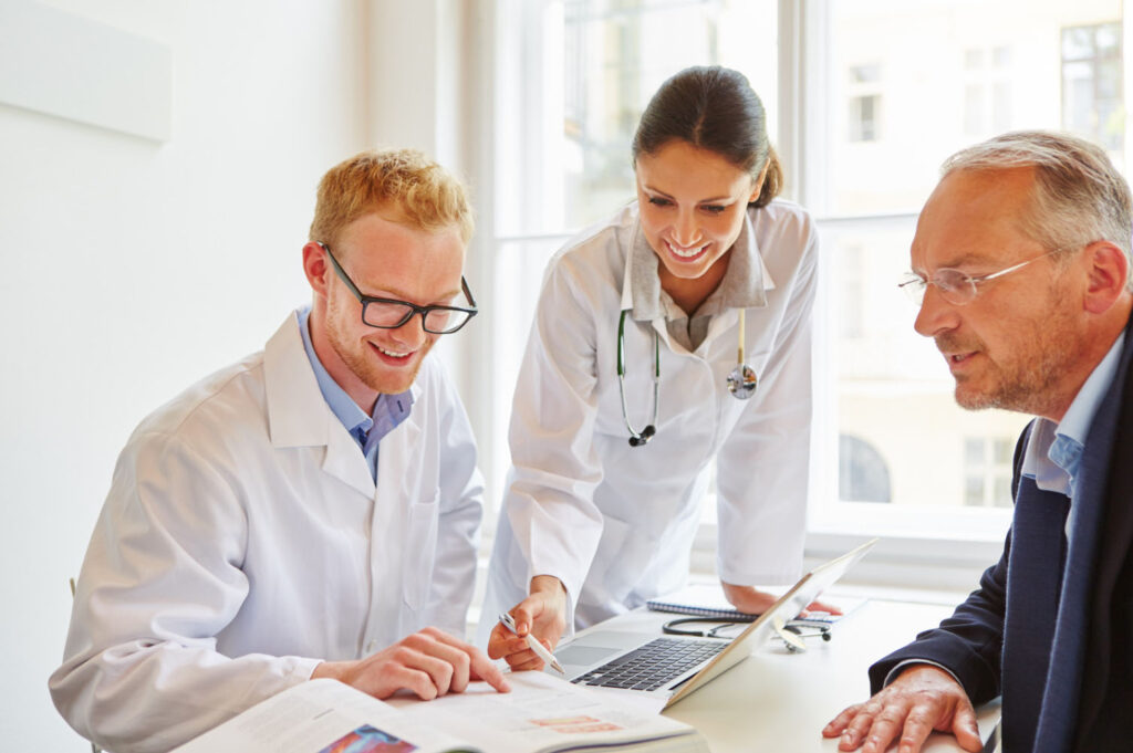 Two doctors in white coats discuss medical information with a patient in an office, smiling and referencing a laptop and medical documents during the consultation.