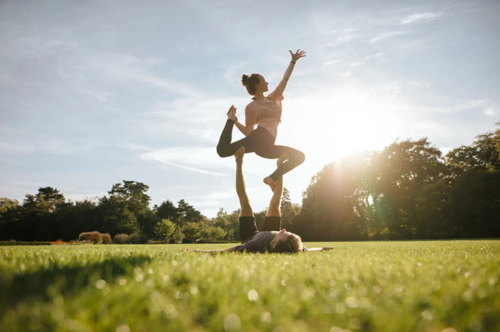 Two women performing an acro yoga pose outdoors in a grassy field with the sun setting in the background. One woman is balancing on the other, with their arms raised towards the sky.