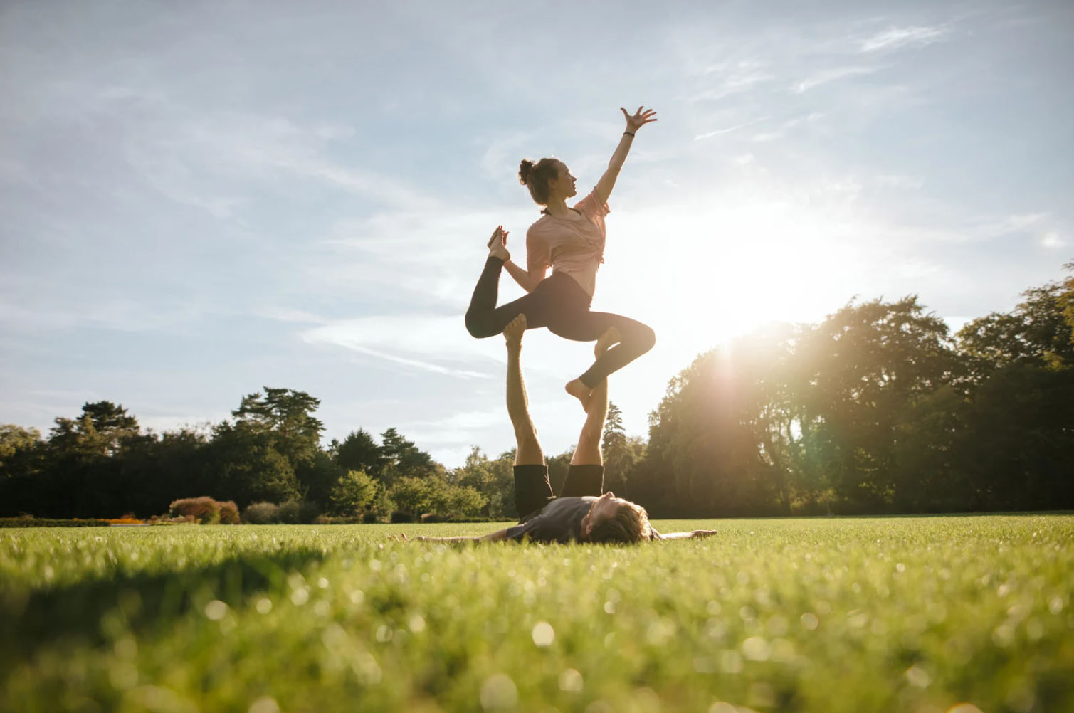 Two women performing an acro yoga pose outdoors in a grassy field with the sun setting in the background. One woman is balancing on the other, with their arms raised towards the sky.