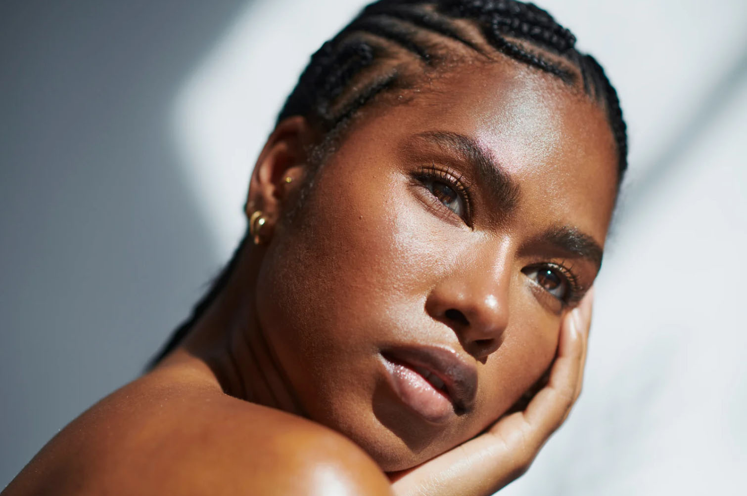 A close-up portrait of a woman with braided hair, showcasing her glowing, radiant skin as she rests her face on her hand, illuminated by soft natural light.