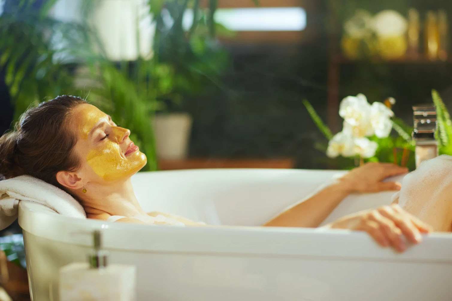 A woman relaxing in a bath with a golden face mask, surrounded by plants and soft lighting, enjoying a moment of self-care.