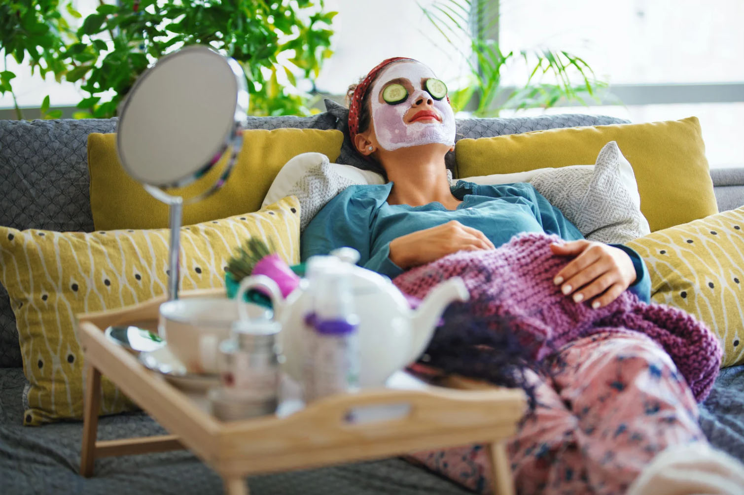 A woman relaxing on a couch with a facial mask and cucumber slices on her eyes, wrapped in a cozy blanket. A tray with tea, skincare products, and a mirror sits beside her, with plants in the background.
