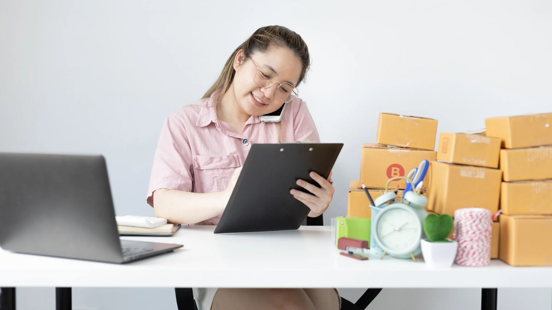 A young woman wearing glasses is multitasking at her desk, talking on the phone while writing on a clipboard. She is surrounded by brown shipping boxes, a laptop, and office supplies, suggesting she is managing an online business or handling e-commerce orders.
