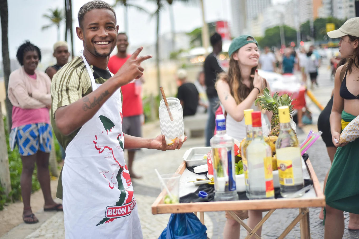 A smiling bartender in an apron serves drinks from a street cart in Rio, with bottles of alcohol and fresh fruits on display, while people enjoy the beachside atmosphere.