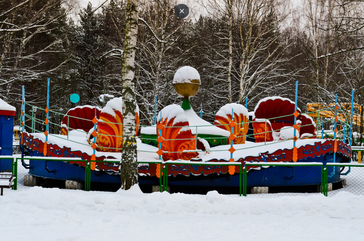 Colorful carousel ride covered in snow at a winter amusement park, surrounded by snow-covered trees and a quiet, serene winter landscape.