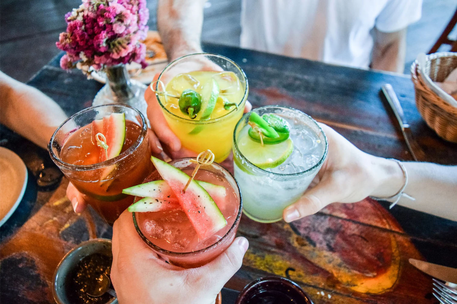 Group of people toasting with colorful cocktails on a wooden table with slices of fruit and herbs as garnish.
